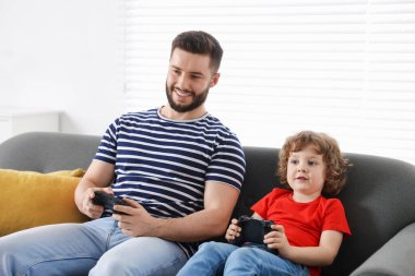 Mutlu baba ve oğlu evdeki kanepede oyun kumandalarıyla oynuyorlar.