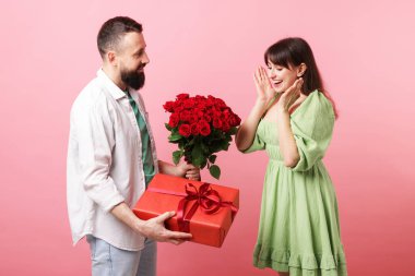 Kırmızı gül buketini ve hediye kutusunu pembe arka plandaki sevgilisine sunan bir adam.