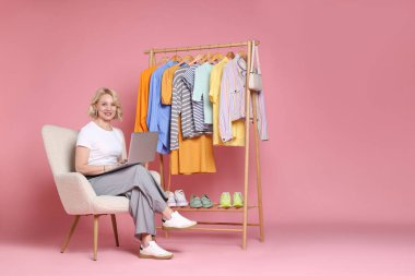 Gülümseyen moda tasarımcısı pembe arka planda dizüstü bilgisayarı ve elbise askısı var.