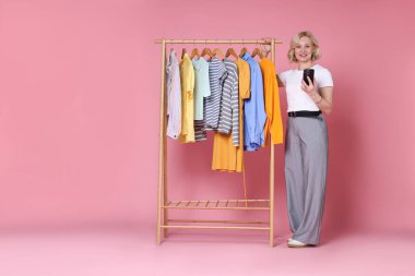 Gülümseyen moda tasarımcısı, akıllı telefonu ve pembe arka planda elbise askısı var. Metin için boşluk