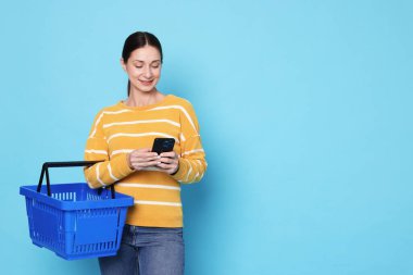 Alış veriş sepeti ve açık mavi arka planda akıllı telefonu olan gülümseyen kadın. Metin için boşluk