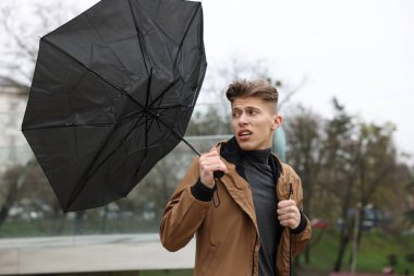 Kırık şemsiyeli bir adam açık havada yağmur altında rüzgarla savaşıyor.