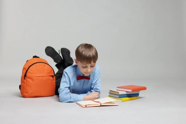 Sırt çantası açık gri arka planda kitap okuyan sevimli bir okul çocuğu.
