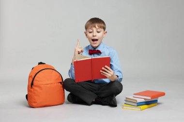 Sırt çantası açık gri arka planda kitap okuyan sevimli bir okul çocuğu.