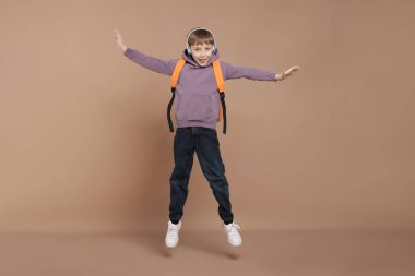 Sırt çantasıyla kahverengi arka planda zıplayan kulaklıklı tatlı bir okul çocuğu.