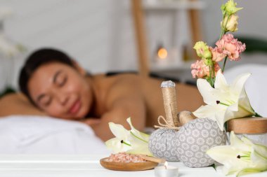 Güzel bir kadın masaj koltuğunda dinleniyor, farklı spa ürünlerine ve zambak çiçeklerine odaklanıyor.