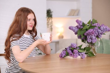 Güzel bir genç kız sıcak bir bardak içecekle ve içeride masada leylak çiçekleriyle