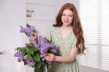 Gülümseyen genç kız evde vazoya leylak çiçekleri koyuyor.