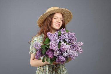 Gri arka planda bir demet leylak çiçeğiyle gülümseyen genç kız.