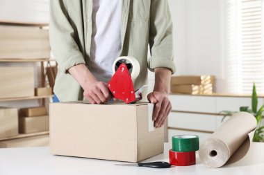 Karton kutuyu kapalı bir yere kapatmak için yapıştırıcılı bant kullanan bir adam.