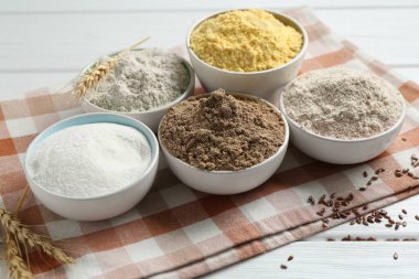 Beyaz ahşap masadaki farklı tipte un, çivi ve keten tohumları, yakın plan.