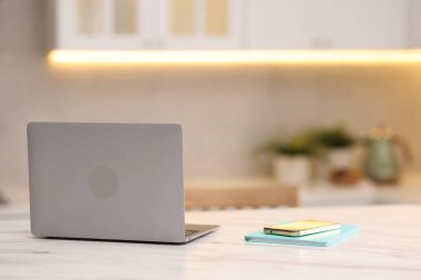 Dizüstü bilgisayar, akıllı telefon ve defter mutfaktaki beyaz mermer masada.