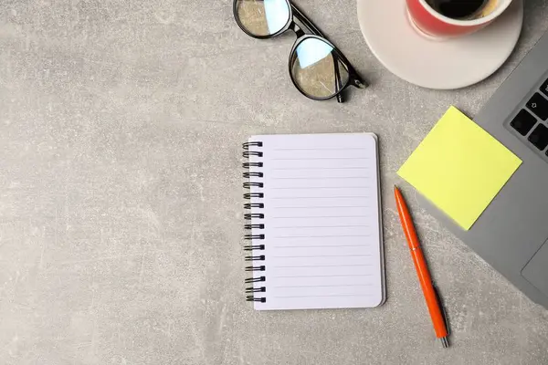 Not defteri, kalem, yapışkan not, bardaklar, kahve ve dizüstü bilgisayar gri masada, düz yerde. Metin için boşluk