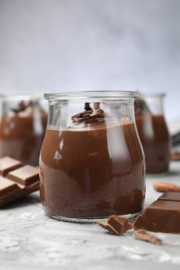 Cam kavanozlarda lezzetli puding ve açık gri masada çikolata parçaları, yakın plan.