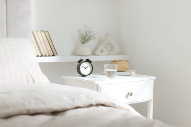 Alarm saati, bir bardak su, hasır kutu ve yatağın yanında kitap olan komodin.