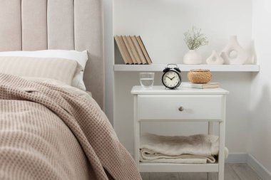 Alarm saati, bir bardak su, hasır kutu ve yatağın yanında kitap olan komodin.