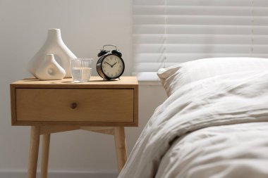 Komodinin yanında çalar saat, bir bardak su ve yatağın yanında vazolar.