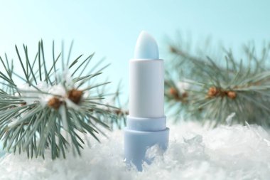 Nemlendirici dudak kremi ve yapay kar üzerindeki köknar ağacı dalları açık mavi arka planda, yakın plan. Kış bakımı