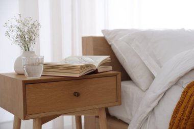 İçinde kitap olan komodin, vazoda çiçekler, yatağın yanında bir bardak su ve bardaklar.