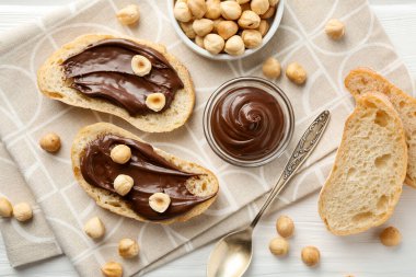 Çikolata sürülmüş ekmek dilimleri ve beyaz ahşap masada fındıklar, düz uzanmış.