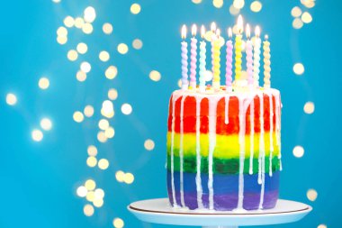 Açık mavi arkaplanda yanan mumlar ve bulanık ışıkları olan enfes bir gökkuşağı doğum günü pastası. Metin için boşluk