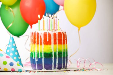 Lezzetli gökkuşağı doğum günü pastası yanan mumlar, flamalar, süslemeler, parti şapkaları ve balonlar beyaz masanın üzerindeki ışık arka planında, yakın plan