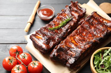 Biberiye, domates, sos ve salata ile servis edilen kızarmış domuz kaburgaları siyah ahşap masada, yakın plan.