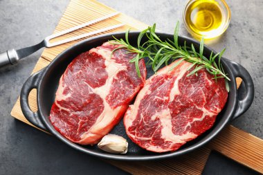 Fırın kabında baharatlı çiğ et parçaları, gri desenli masa üzerinde yağ ve çatal, düz yatar.