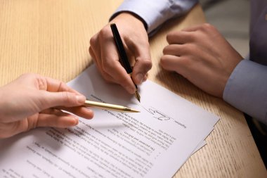 Kadın belgeye işaret ediyor ve adam ahşap masaya imza atıyor, yakın plan.