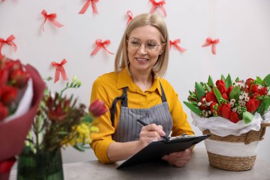 Elinde pano olan bir işyeri sahibi çiçekçi dükkanındaki masada notlar alıyor.