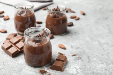 Cam kavanozlarda lezzetli puding, çikolata parçaları ve açık gri masada kakao çekirdekleri, yakın plan. Metin için boşluk