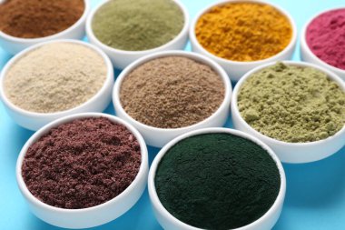 Açık mavi arka planda kaselerde farklı süper yiyecek tozları, yakın plan.