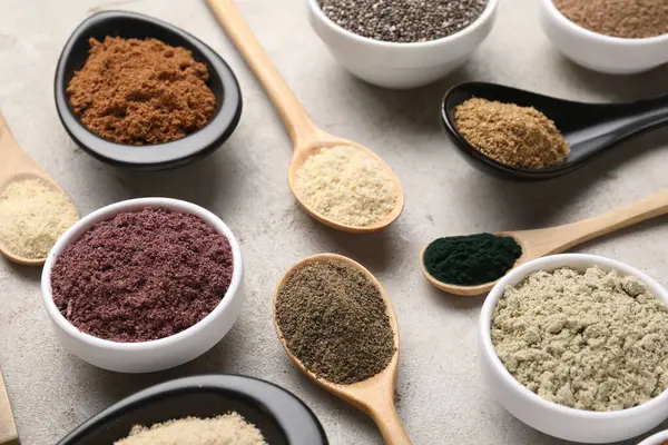 Gri desenli masa üzerinde kaşıklar ve kaseler halinde farklı süper yiyecek tozları.