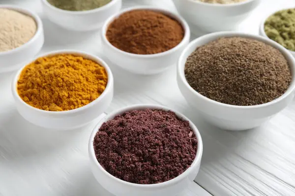 Beyaz ahşap masadaki kaselerde farklı süper yiyecek tozları, yakın plan.