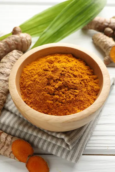 Aromatik zerdeçal tozu, taze kökler ve beyaz ahşap masa üzerindeki yapraklar, yakın plan.