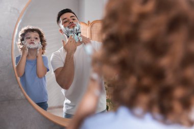 Banyodaki aynanın yanında yüzlerinde tıraş kremi olan mutlu baba ve oğlu, seçici bir odak noktası.