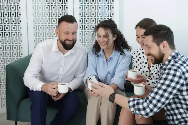 Ofisteki kahve molasında iş arkadaşlarına telefonunda bir şey gösteren bir adam.