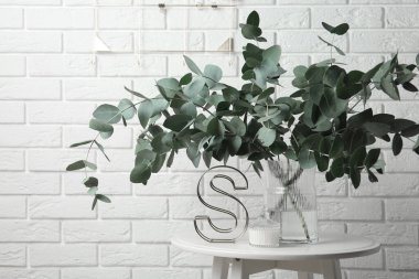 Vazoda yeşil okaliptüs dalları ve beyaz tuğla duvarın yanındaki masada diğer dekorlar.
