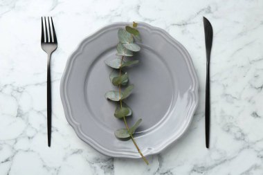 Beyaz mermer masa üzerinde şık bir tabak, çatal bıçak ve okaliptüs dalı.