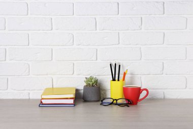 Farklı kırtasiye malzemeleri, kahve, bardak ve çiçek dekoru beyaz tuğla duvarın yanındaki ahşap ofis masasında.