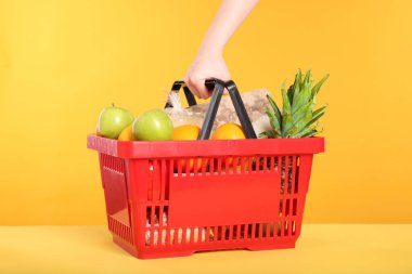 Sarı arka planda ürünlerle dolu alışveriş sepeti olan bir kadın.