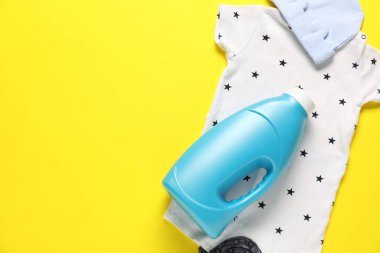 Bir şişe yumuşatıcı ve sarı arka planda bebek kıyafetleri. Metin için boşluk