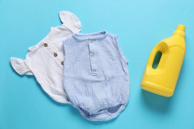 Bir şişe yumuşatıcı ve açık mavi arka planda bebek kıyafetleri.