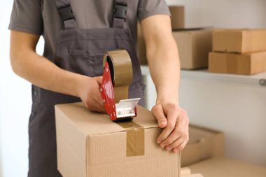 Karton kutuyu kapalı bir yere kapatmak için yapıştırıcılı bant kullanan bir adam.