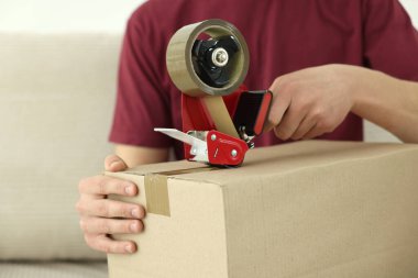 Karton kutuyu kapalı bir yere kapatmak için yapıştırıcılı bant kullanan bir adam.