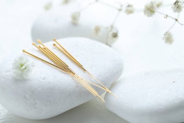 Akupunktur iğneleri, çakıl taşı ve çiçek tomurcukları beyaz desenli masada, yakın plan.