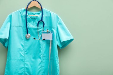 Steteskop ve rozeti açık yeşil arka planda asılı sağlık görevlisi üniforması. Metin için boşluk