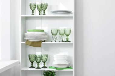 Beyaz duvarın yanındaki raflarda temiz bardaklar ve tabaklar var.