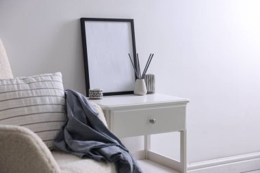 Boş çerçeve, sazlık dağıtıcı, koltuk ve şık ev dekorasyonu beyaz duvarın yanındaki masada.