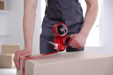 Karton kutuyu kapalı bir yere kapatmak için yapıştırıcılı bant kullanan bir adam.